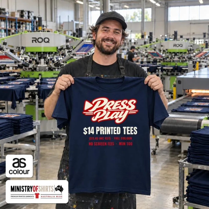 Printer holding up a navy Press Play tee in front of automatic screen printing presses